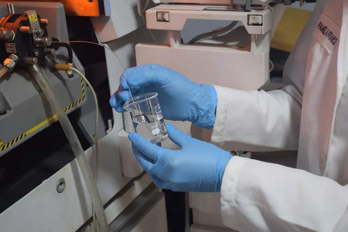 Technician in blue gloves holds water sample in test tube at laboratory bench with analytical equipment in background