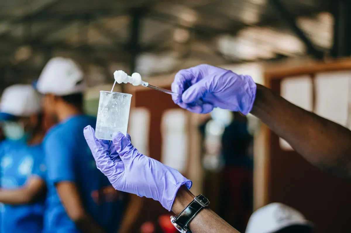 Technician in purple gloves holds clear water sample in glass container during borehole yield test, with workers and hard hats visible in background