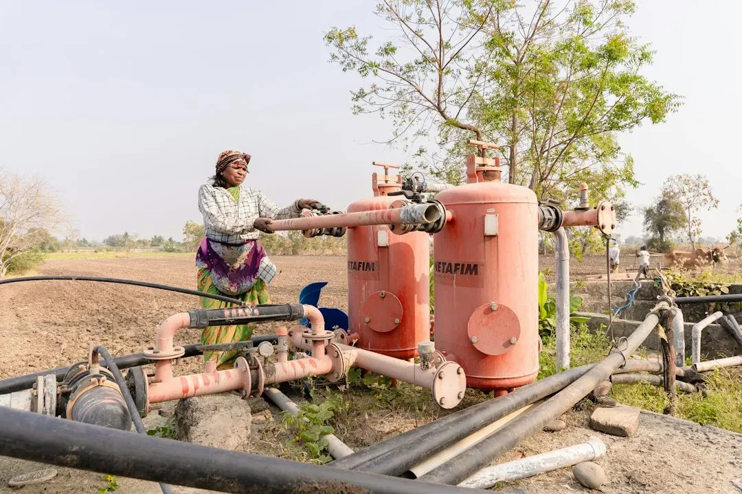 Woman operating borehole water pumping system with red pressure tanks and piping in rural setting
