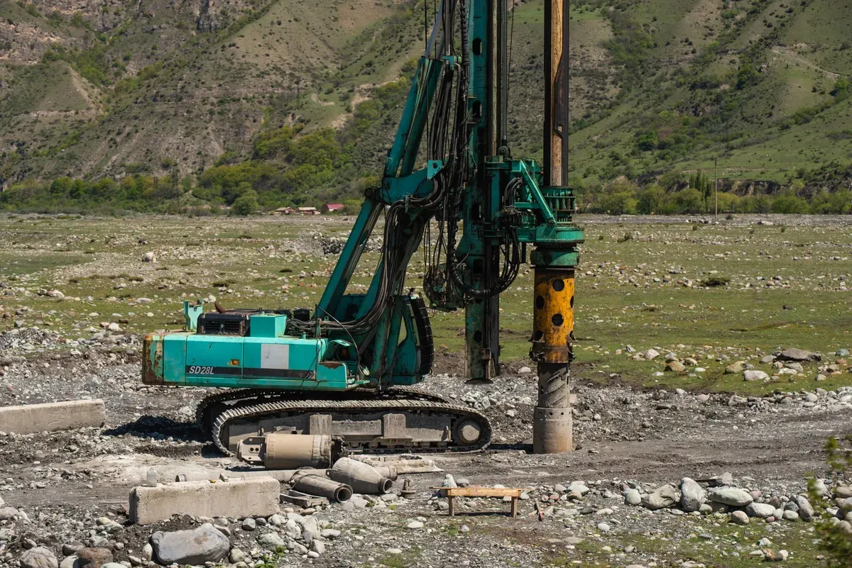 Teal drilling rig with mast mounted on excavator, drilling borehole with casing pipes and collar visible, in mountainous valley landscape