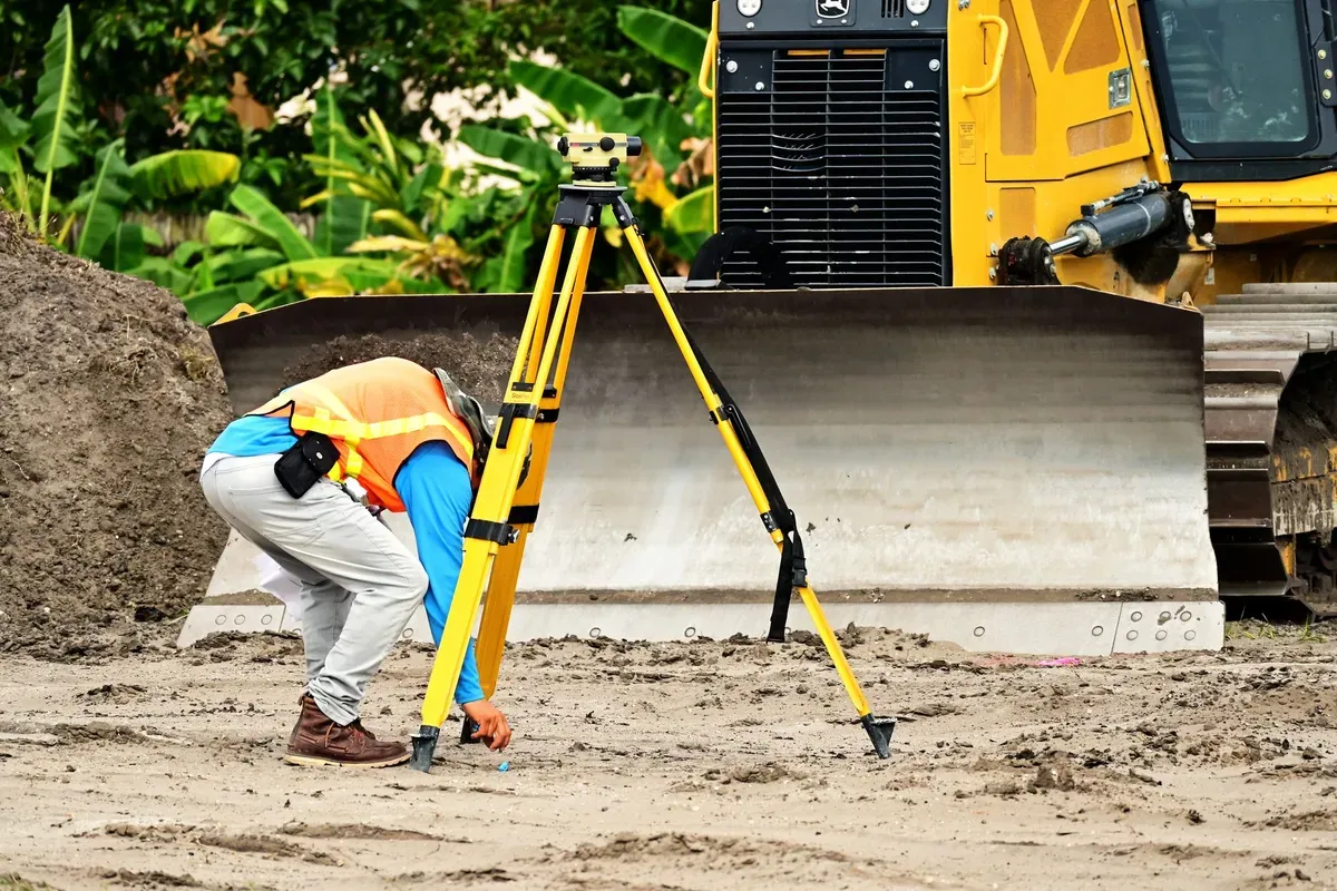 Surveyor using tripod-mounted theodolite to conduct geophysical borehole site assessment with survey vehicle behind